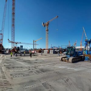 a construction site bustling with workers and machinery, surrounded by towering cranes under a clear blue sky.