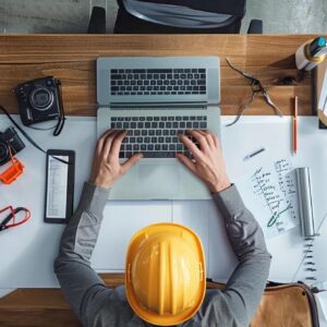 Person wearing a yellow hard hat typing on a laptop at a construction desk with tools, notepad, and camera, illustrating digital marketing strategies for construction firms.