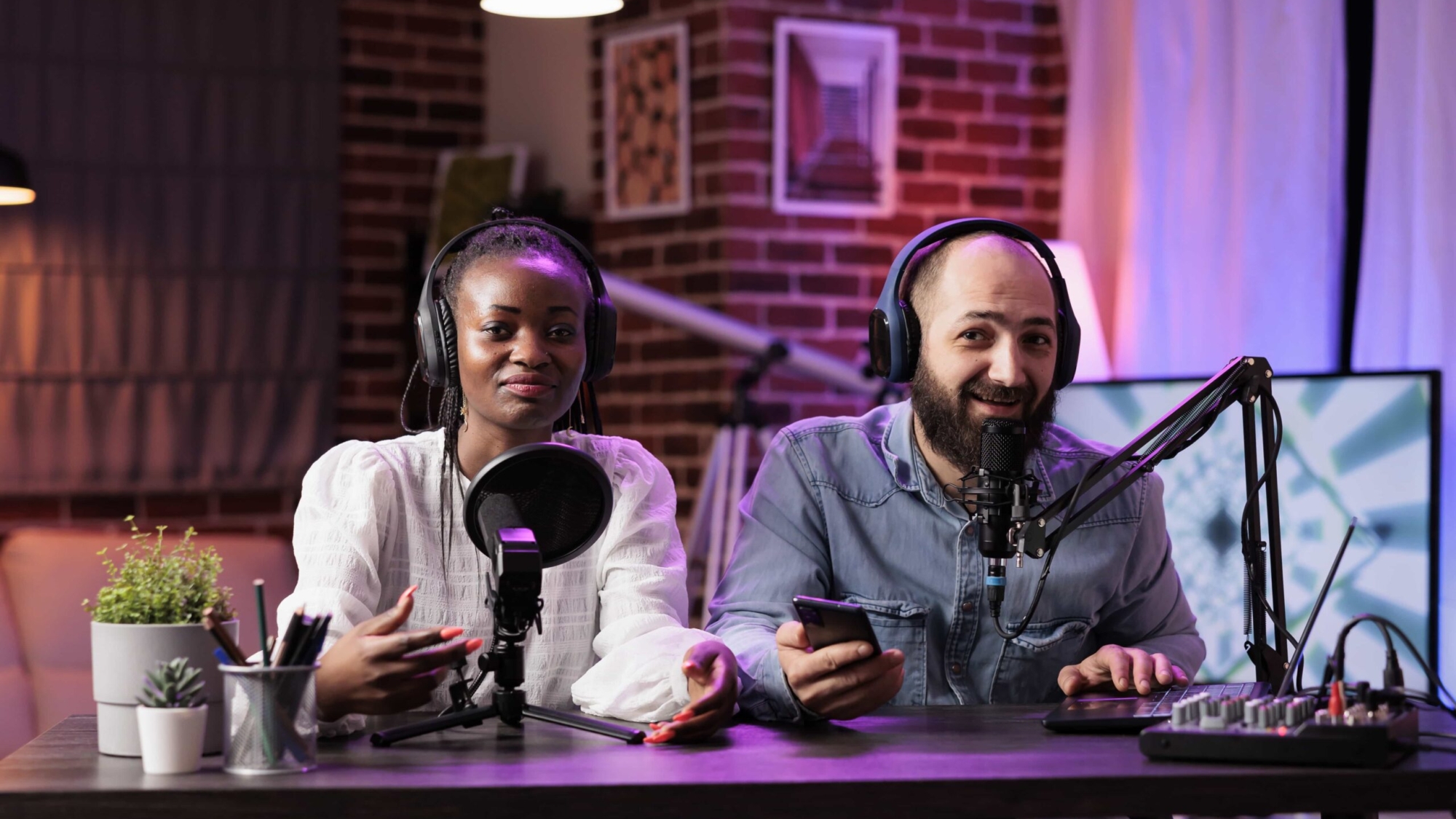 Podcasters discussing construction topics, featuring a woman with a microphone and a man holding a smartphone, in a studio setting with recording equipment and a modern backdrop.