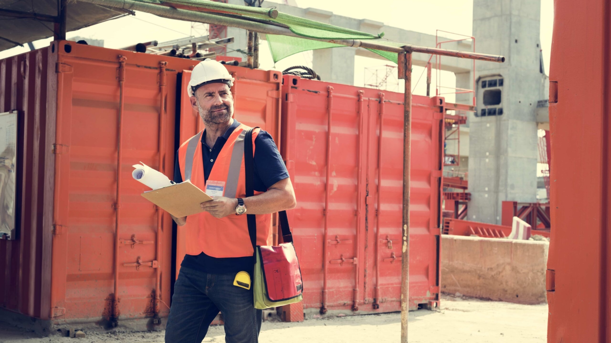 Construction executive in safety gear holding blueprints and clipboard, standing near orange shipping containers on a construction site, illustrating the application of StoryBrand principles in commercial construction marketing.