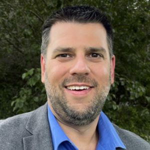 Perryn Olson smiling in a professional setting, wearing a blue shirt and gray blazer, discussing branding strategies for construction firms to attract clients and talent.