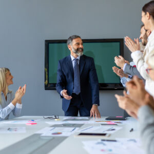 Business meeting with a smiling man in a suit standing at a table, receiving applause from colleagues, symbolizing effective leadership and collaboration in marketing strategies for construction businesses.