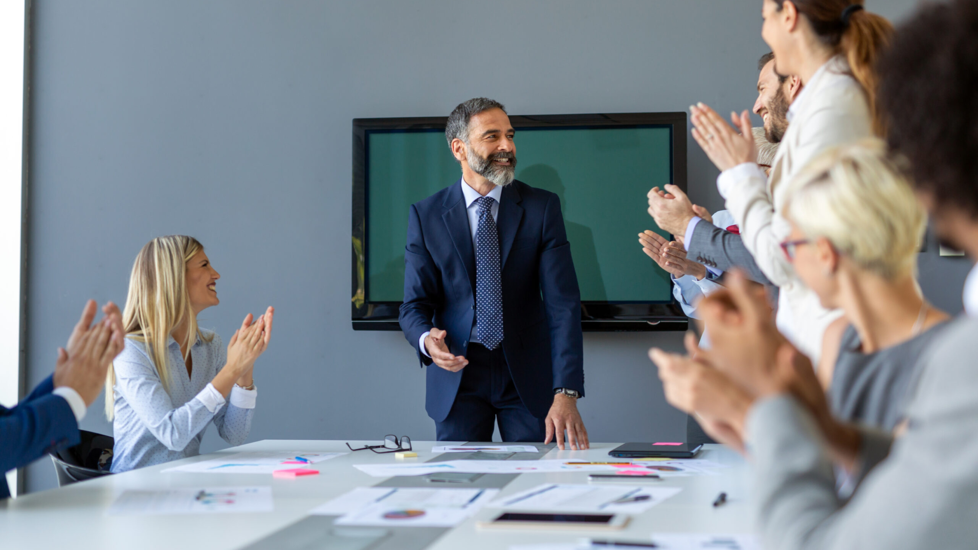 Business meeting celebrating success, with a smiling man in a suit receiving applause from colleagues around a table covered in documents and notes, emphasizing teamwork and leadership in marketing strategies.