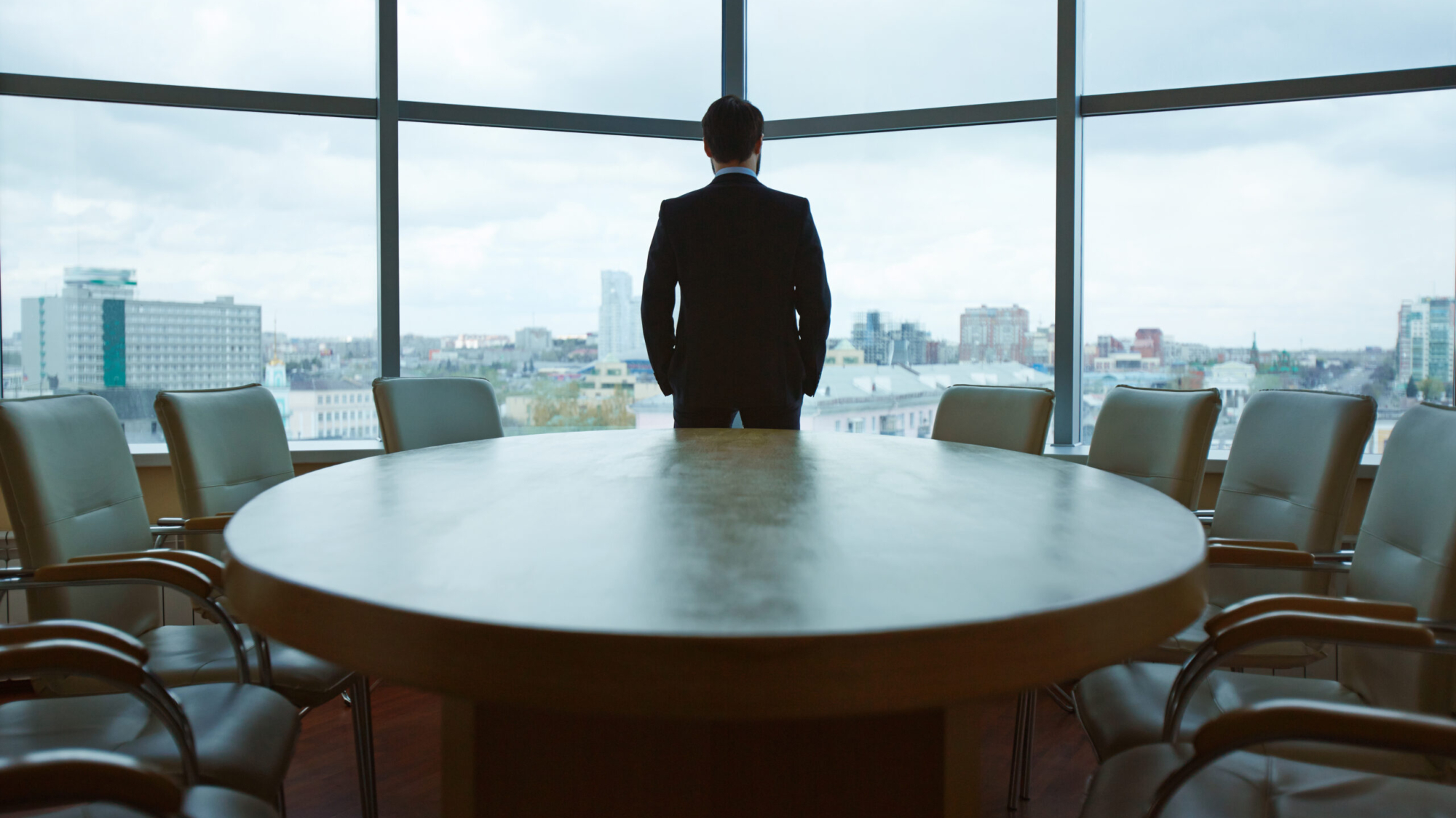 Business professional in suit standing by large conference table, gazing out at city skyline, symbolizing strategic leadership and career advancement in marketing.
