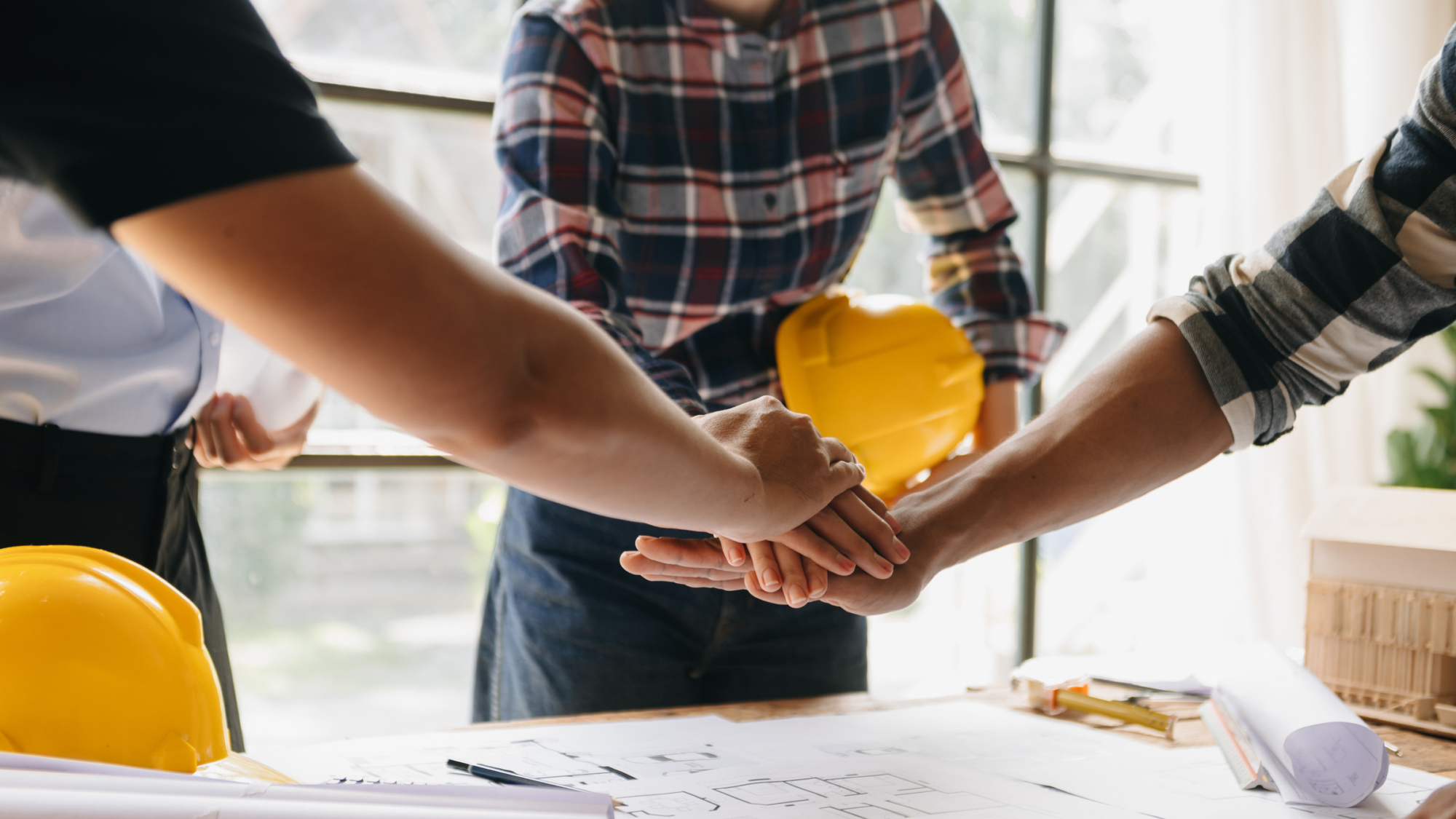 Hands joining in collaboration over construction plans, with a yellow hard hat in the background, symbolizing teamwork in the construction industry.