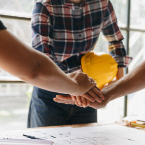 Hands of construction professionals collaborating over project plans, with a yellow hard hat in the background, emphasizing teamwork in construction marketing strategies.